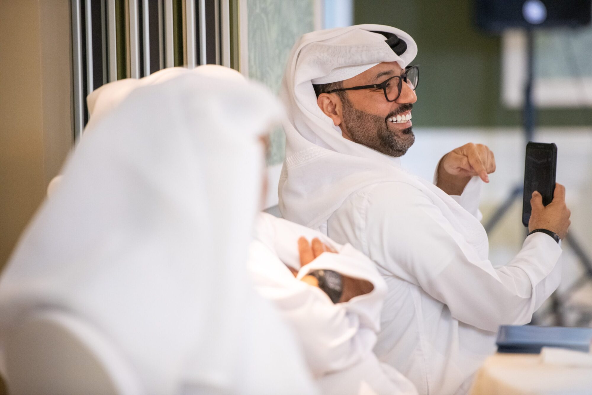 Two Qatari men laughing, one holding a phone and pointing at it.