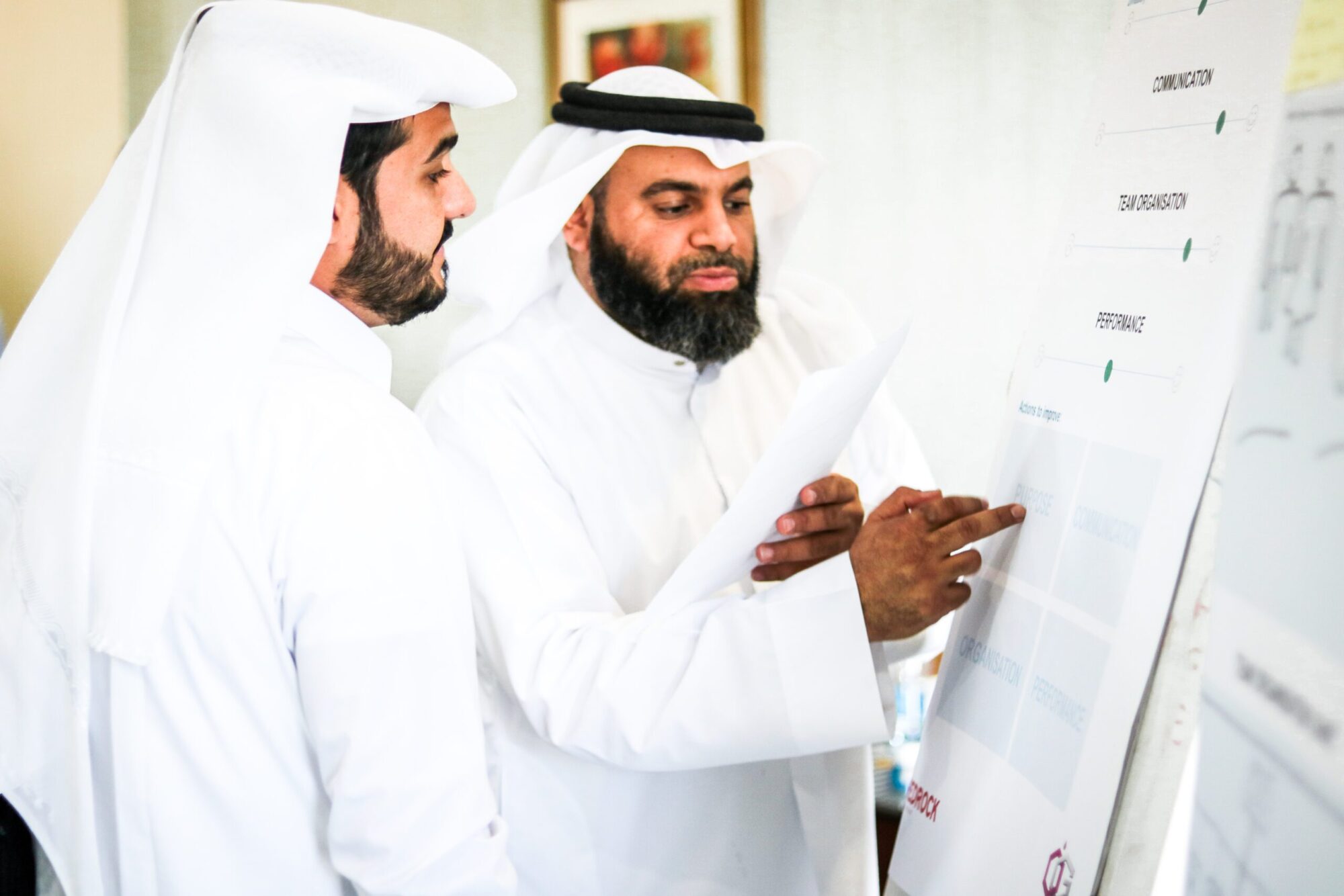 Two Qatari men standing in front of a whiteboard, talking and pointing at it.