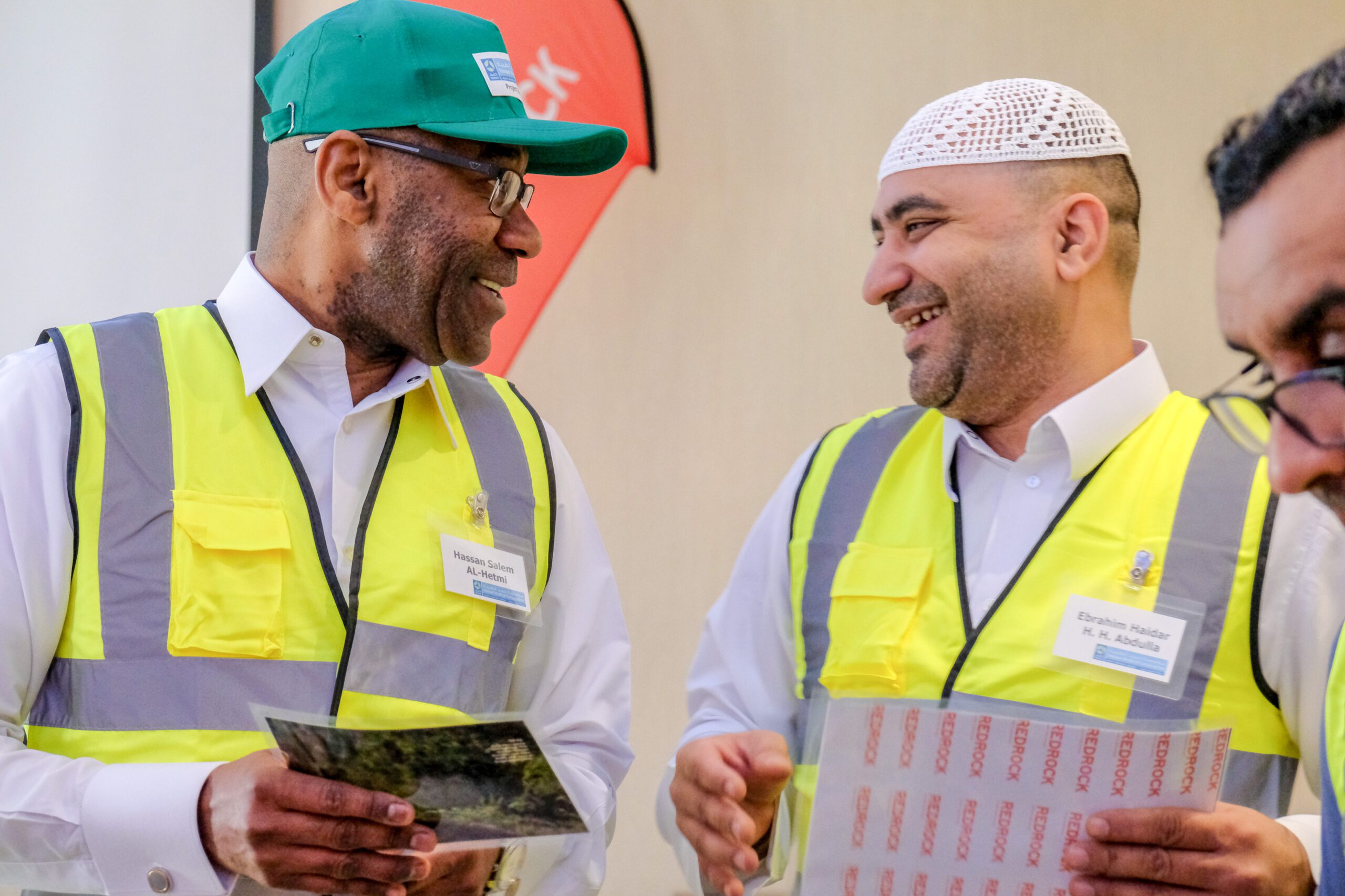 Two men in high-visibility jackets smiling at each other while holding clipboards.