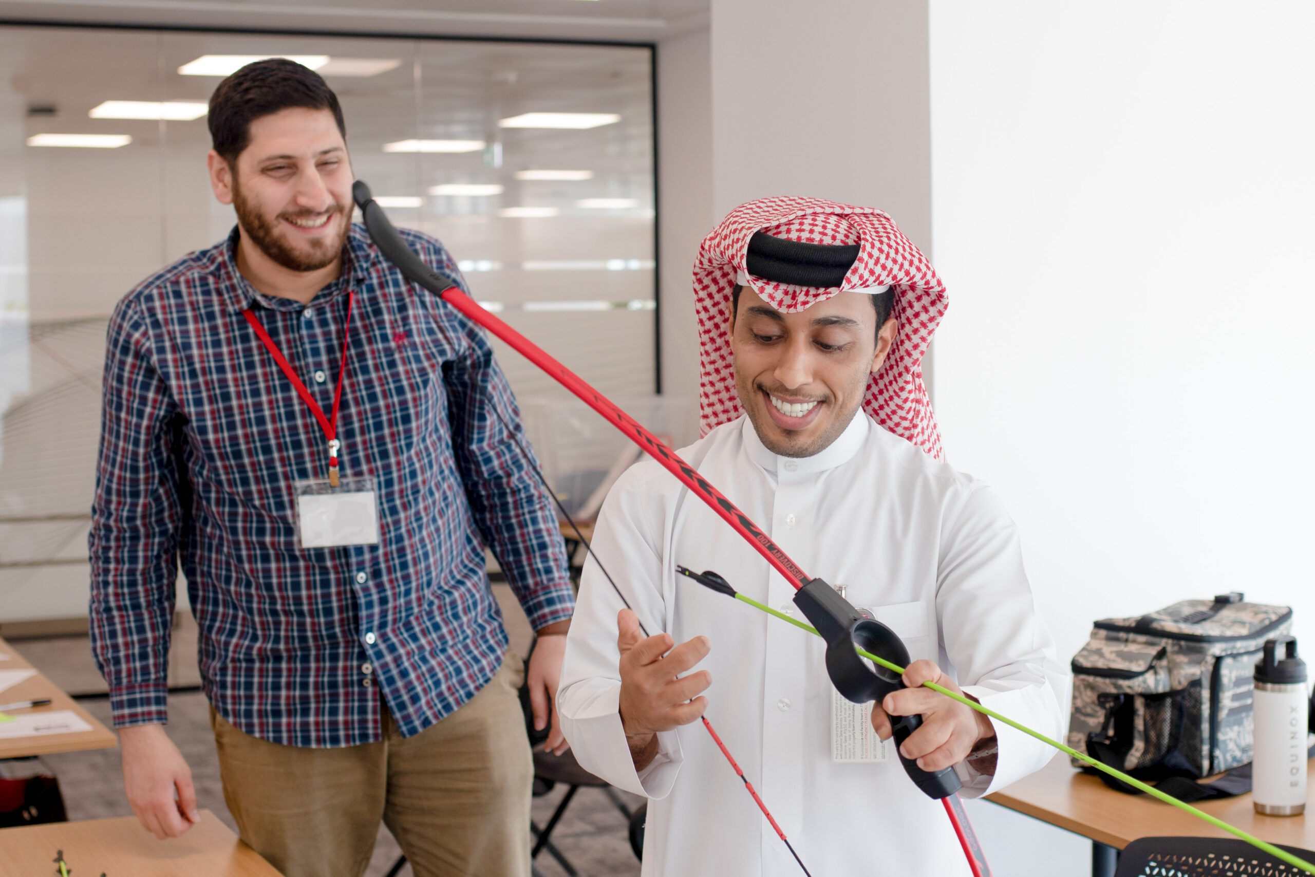 Man prepares to load bow and arrow toy while another man looks at him while smiling.