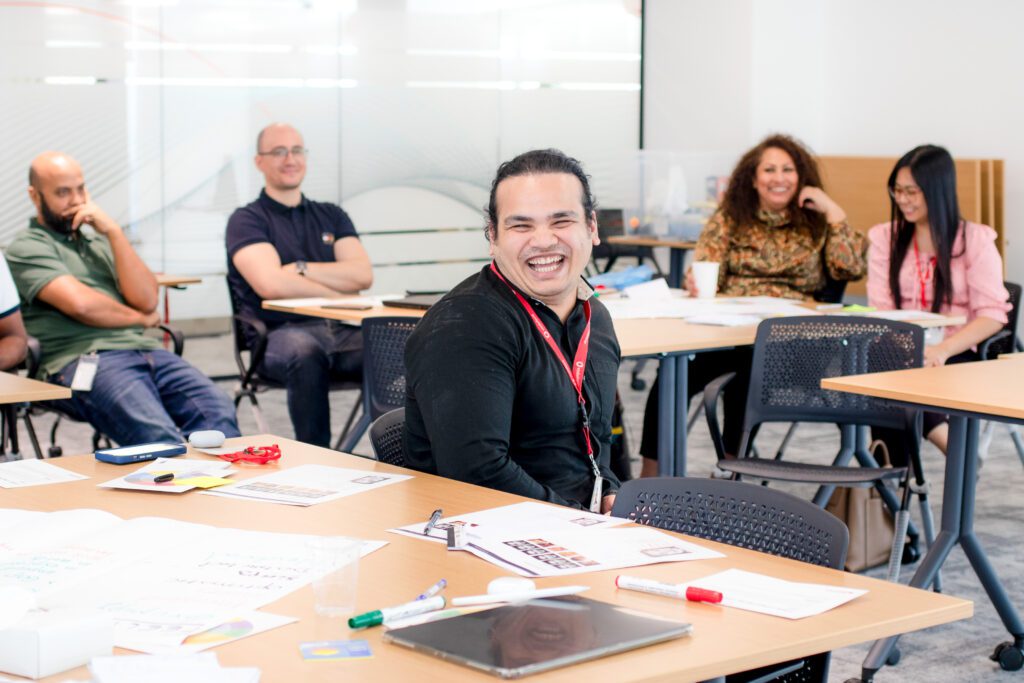 A man doing a big smile at the camera while sat in a classroom environment.